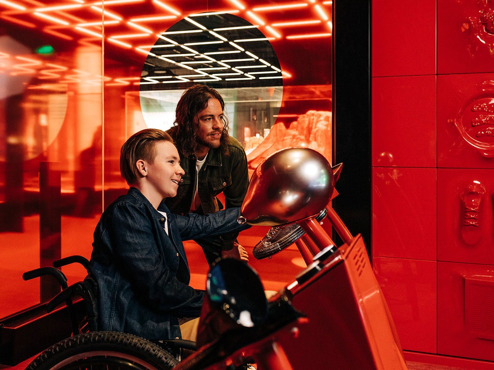A man crouching and a boy in a wheelchair play with silver objects in a red room