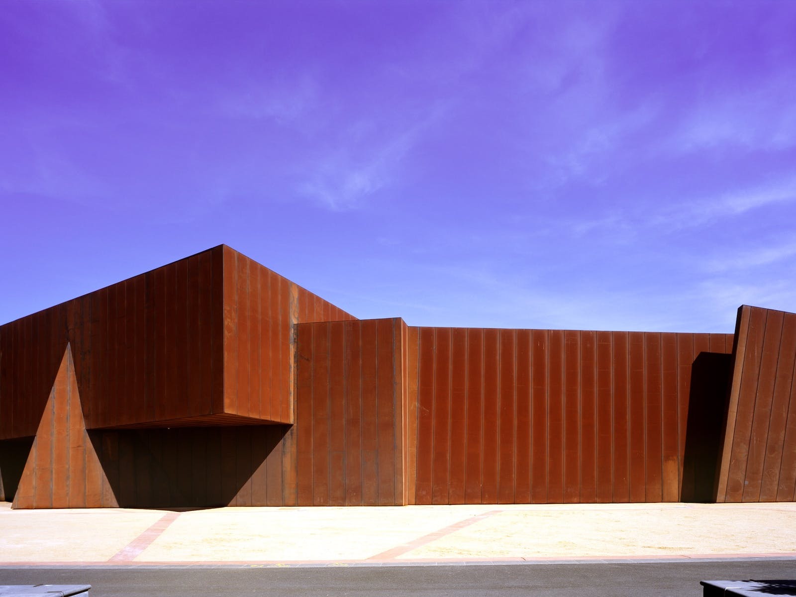 Image of a rust coloured building with sandy gravel in the foreground and a blue sky overhead