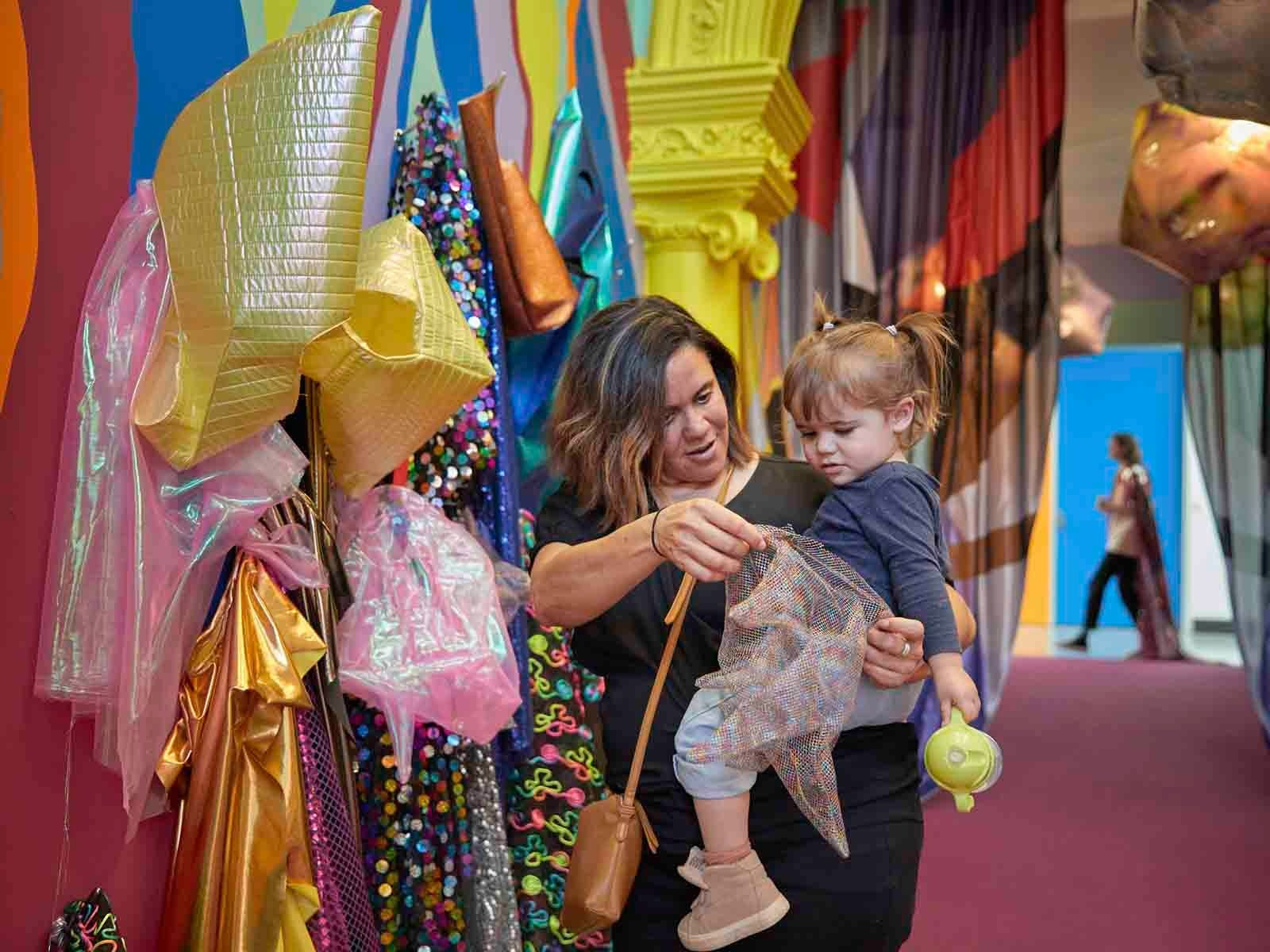 Mother and toddler look at glittering costumes in a brightly painted gallery