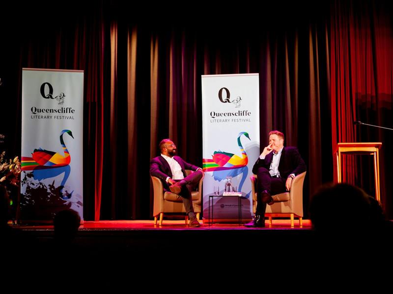 Two men sit on stage; there are QLF banners behind them