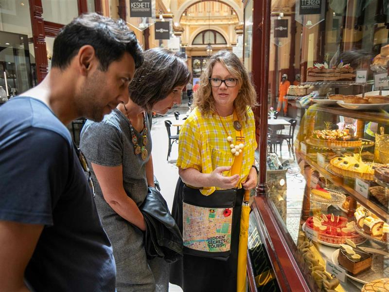 Guide wearing a branded tour bag and yellow top, points oto cakes in a window to 2 tour guests
